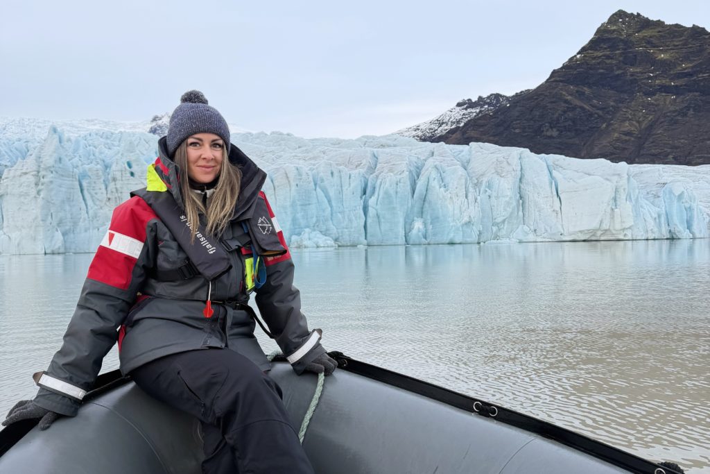 Team member Livia sitting on a small boat in front of a glacier, dressed in cold-weather gear and smiling,