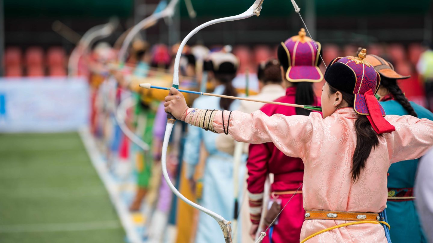 A row of people in vibrant costumes with hats and bows, ready to shoot arrows in an outdoor event.