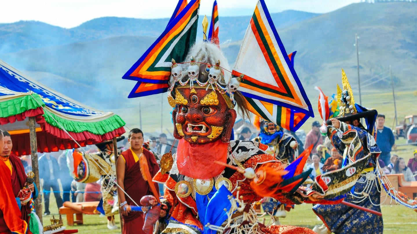 Performers in colorful and intricate costumes, including masks and headpieces, participate in a traditional dance.
