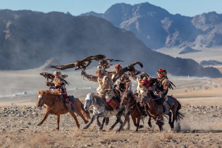 A group of horse riders in a desert, holding large birds of prey with wings open.