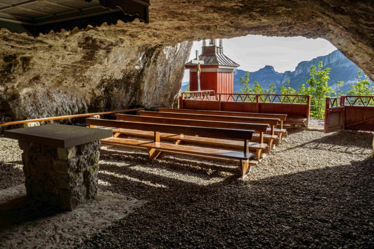 A unique sanctuary within a cave, with wooden benches, a stone altar, the opening shows a scenic view of greenery and mountains.