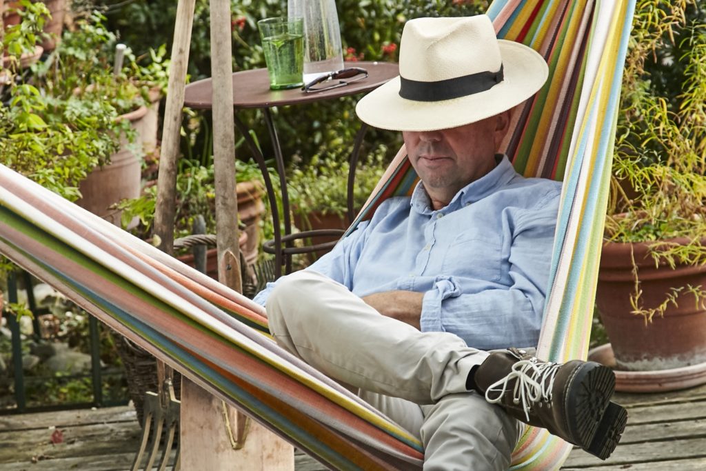 A colorful hammock with a person wearing a hat resting in a green garden with potted plants and a drink on a table.