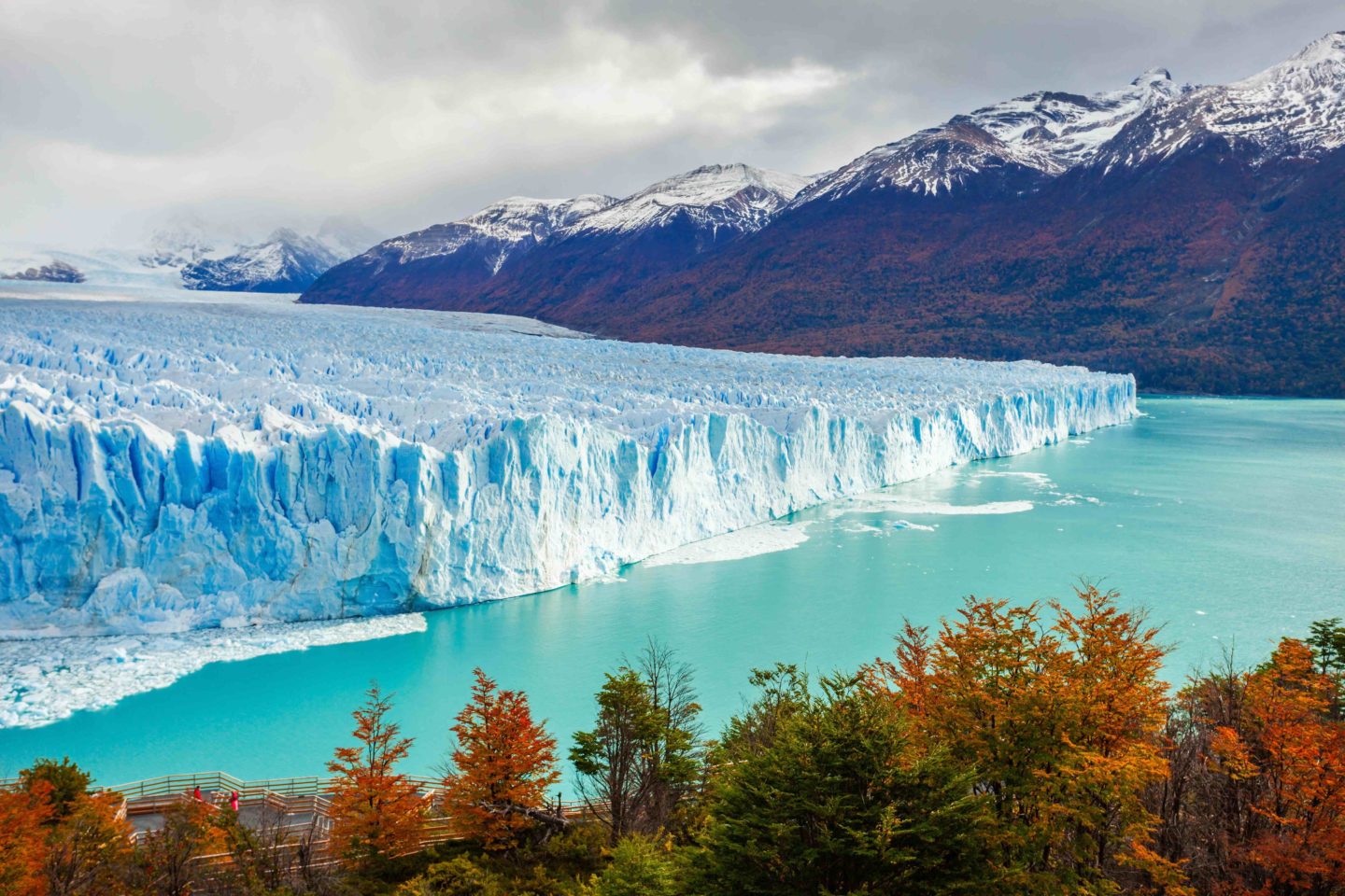 A turquoise glacier with water around it and autumnal trees in the foreground.