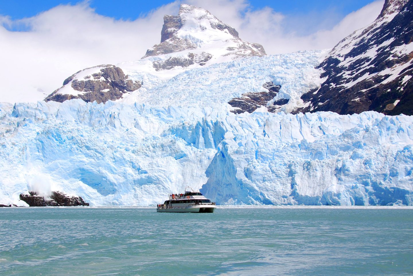 Boat cruising on calm waters, surrounded by blue glacier that extends from the base of snow-covered mountains.
