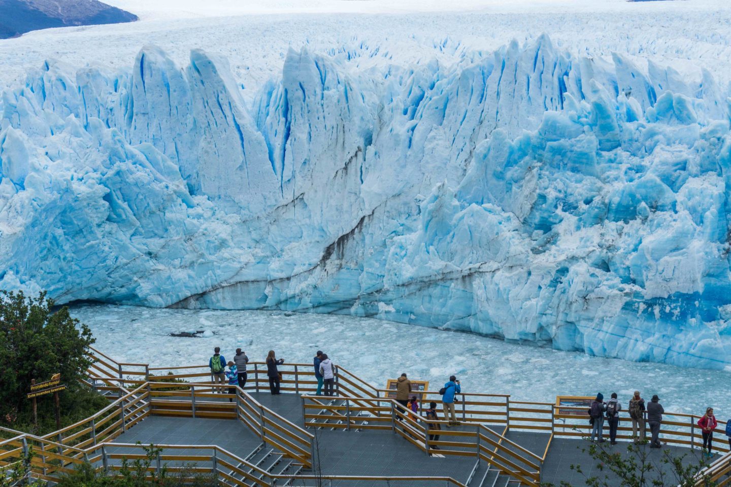 Tourists on a wooden platform with railings, looking at a blue glacier.