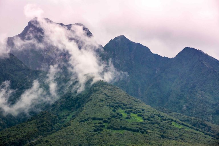 A scenic view of a green mountain range with white clouds covering the peaks and a blue sky above.