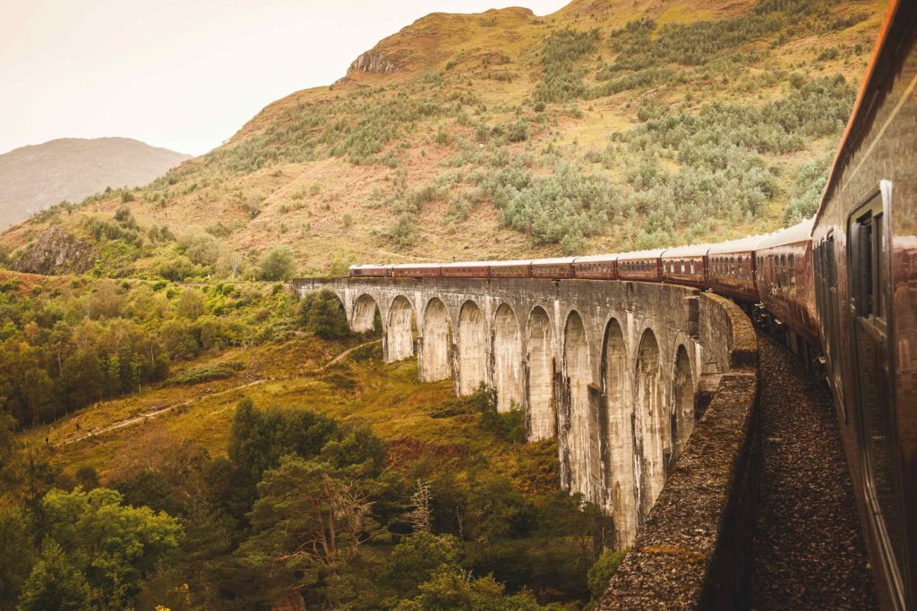 A train crossing a stone bridge with arches, surrounded by greenery and hills.