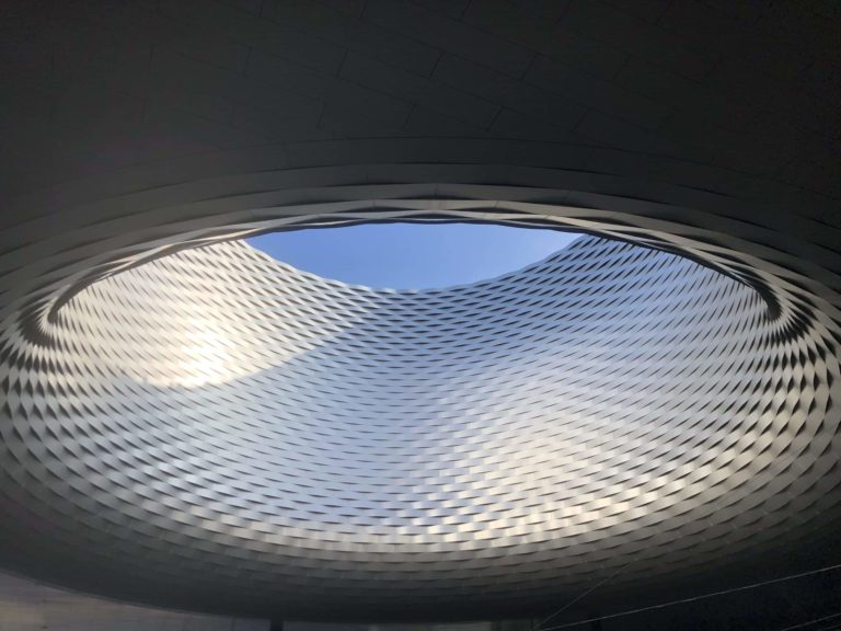 An architectural masterpiece showcasing a circular opening revealing the clear blue sky, surrounded by a patterned structure creating wave-like illusions.