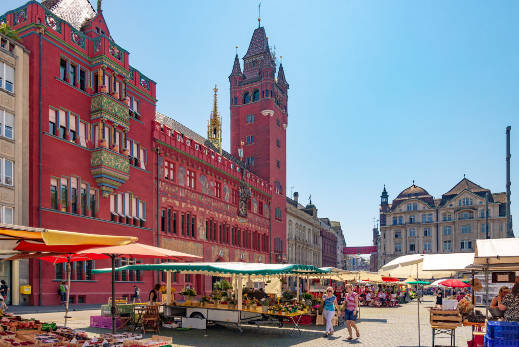 A vibrant market scene in Marktplatz with a prominent red historic building and tower in the background, under a clear blue sky.