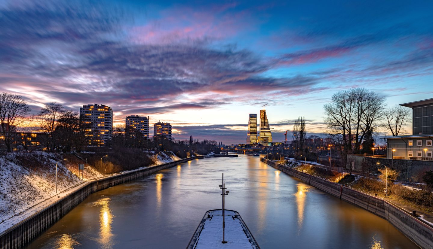 A serene view of a cityscape during sunset, with the sky painted in hues of blue, pink, and orange. Buildings silhouette against the vibrant sky, and their lights reflect on the calm Rhine river below.