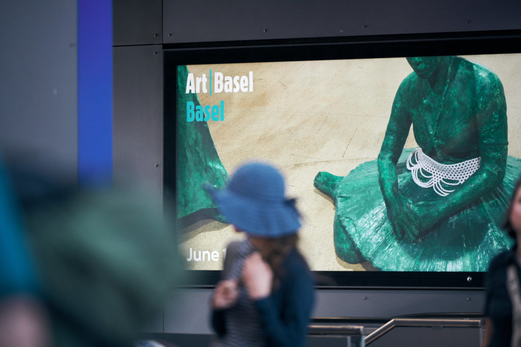 A lively street scene with a person walking by an Art Basel advertisement showcasing a green sculpture.