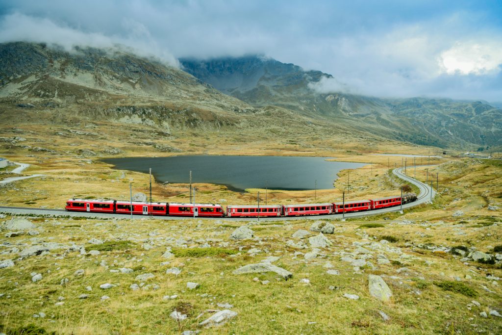 Bernina Railway traversing a mountainous landscape, with a serene lake in the background, showcasing the beauty of the Swiss Alps.