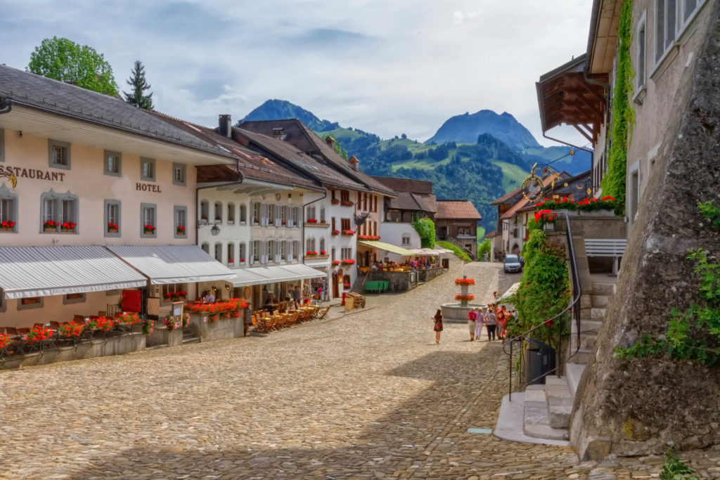 Picturesque view of a cobblestone street in a mountain village, with traditional alpine architecture, colorful flowers, and distant green mountains.