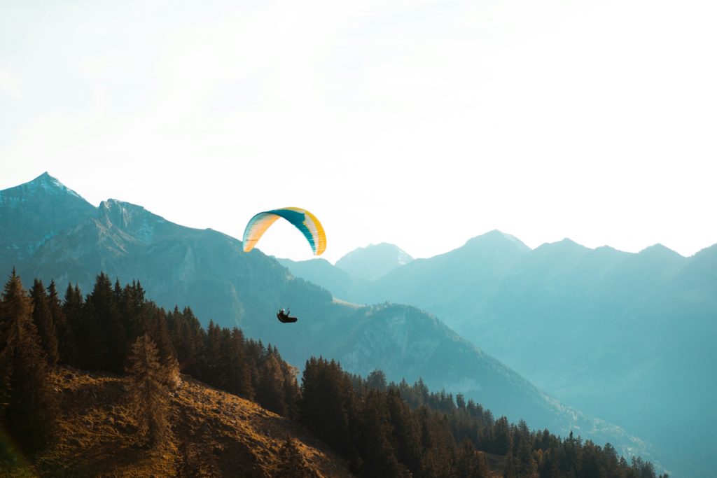 A paraglider soars over forested mountains, with the peaks highlighted by the golden hue of the sun.