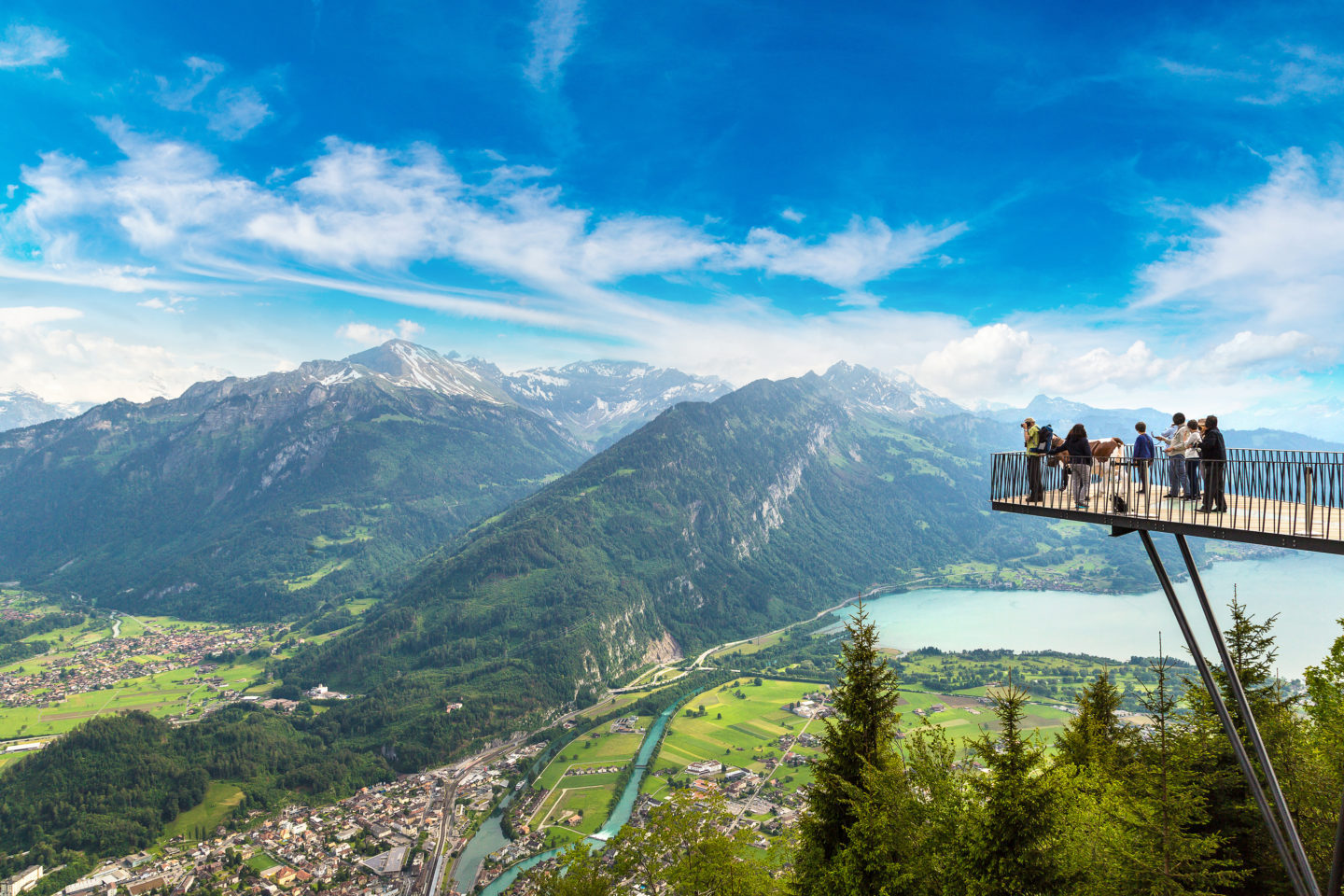 Visitors on a viewing platform overlooking a scenic landscape of mountains, a winding river, and lush forests.