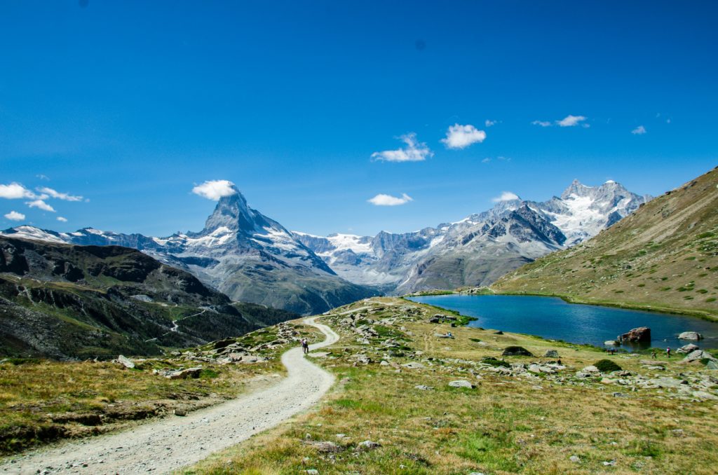 A winding trail leading towards the iconic Matterhorn mountain, with a mountain lake in the foreground and a backdrop of alpine scenery under a bright blue sky.