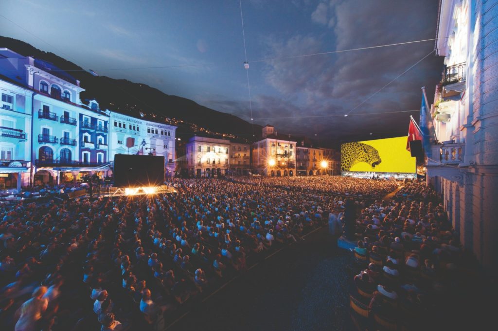 Locarno Film Festival: A bustling large square filled with people seated, captivated, and watching a screen in the enchanting evening atmosphere.
