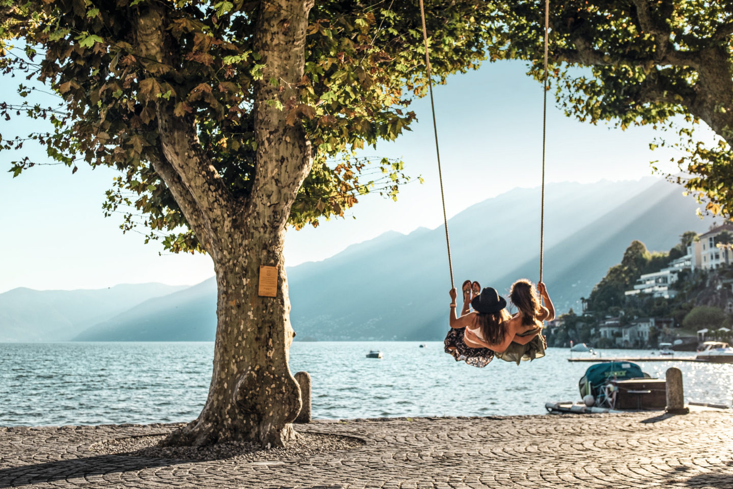 Two young women seated on a swing, joyfully swinging across the lake into the warm embrace of the sunset, creating a magical and carefree moment.