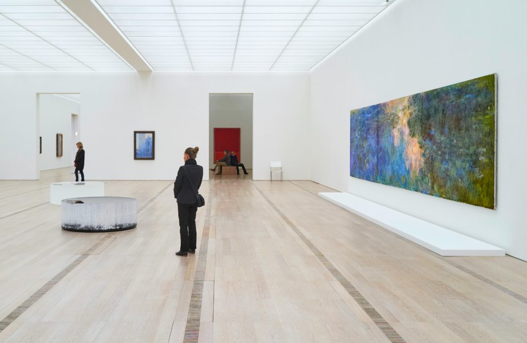 A person observing a large, colorful abstract painting in a bright, spacious art gallery.