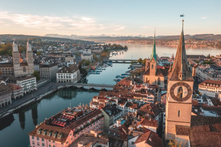 View of Zurich at sunrise, showcasing the city’s historic architecture, river, and distant mountains.