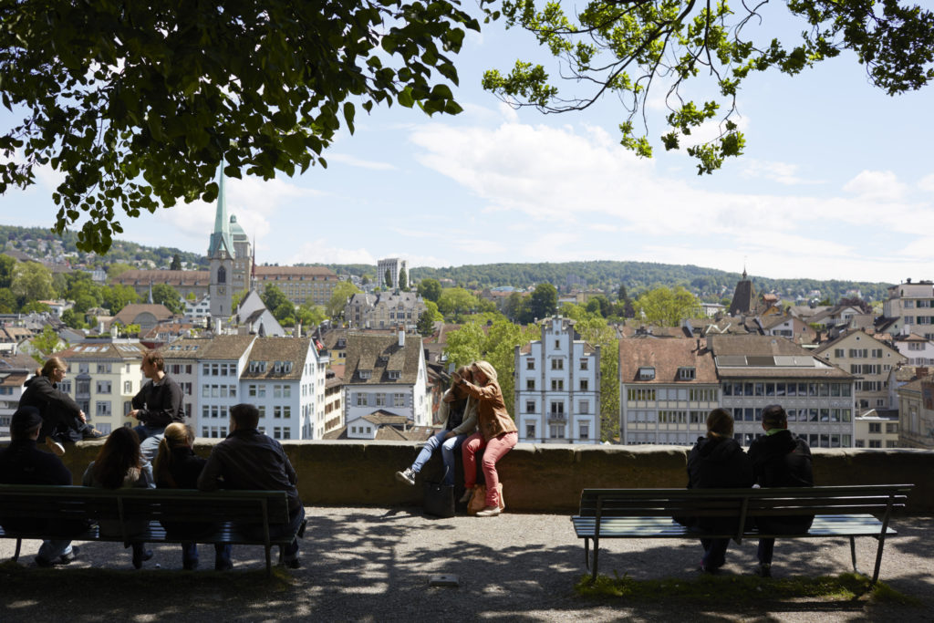 People enjoying a panoramic view of Zurich city with historical architecture from a shaded viewpoint.