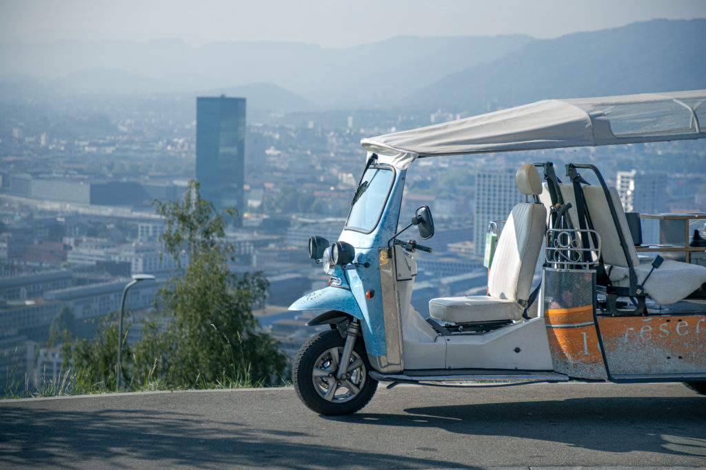 A tuk tuk parked on a hill with a panoramic view of the city in the background.