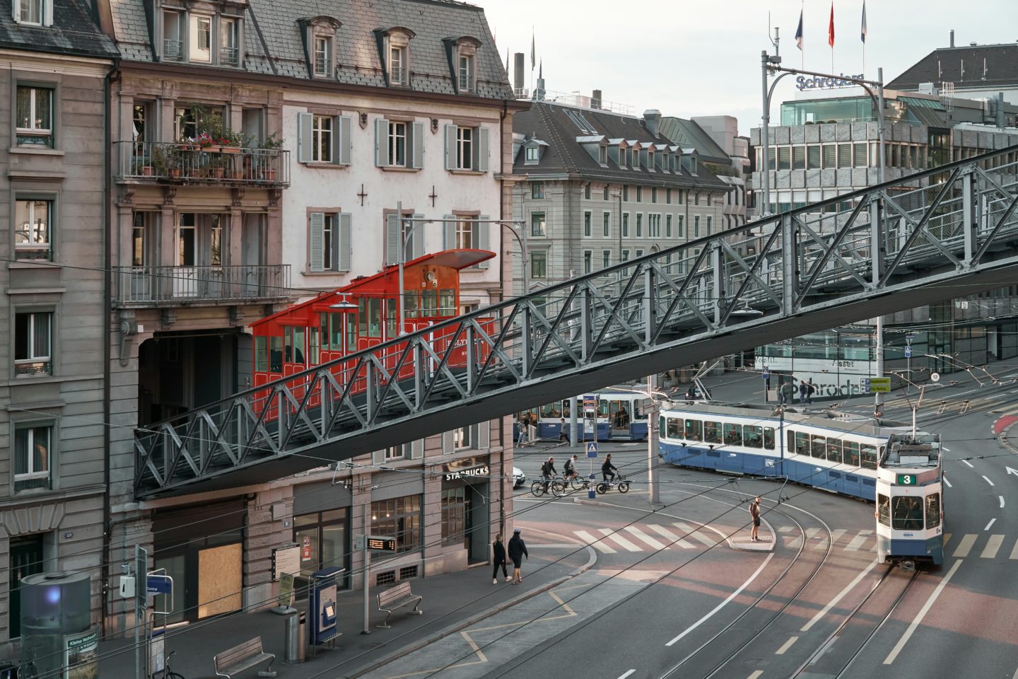 A tram moving through a city intersection beneath a cable car pedestrian overpass