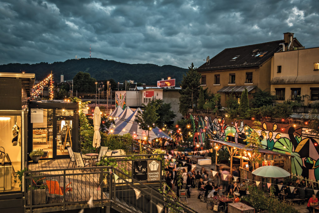 People enjoying their time in an outdoor setting surrounded by artistic walls, under the soft glow of lights with the cityscape in the background.