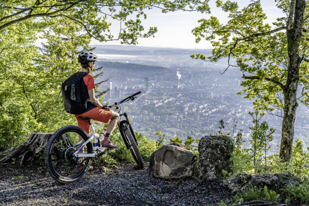 A person on a mountain bike pauses at a high vantage point, overlooking a sprawling city below, surrounded by lush greenery.