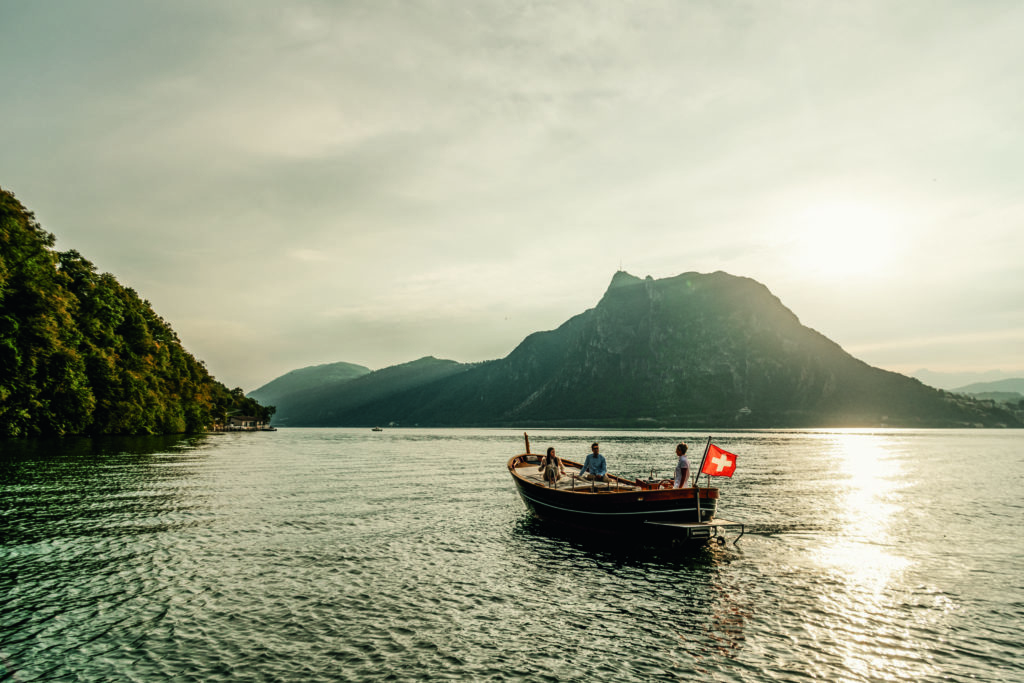 Scenic view of a boat cruising on Lake Lugano proudly displaying the Swiss flag, capturing the tranquil beauty of the Swiss landscape.