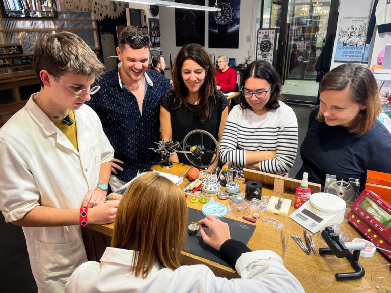 Group gathered around a table observing a woman diligently working on a watch