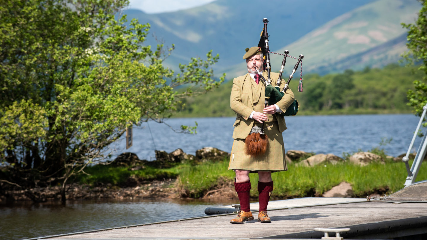 A bagpipe plays on the banks of Loch Lomond in a kilt at an event in Scotland.