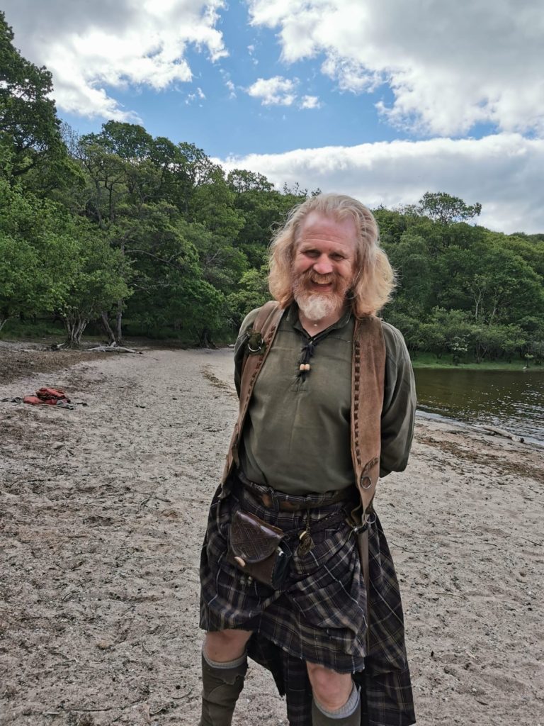 A man in a kilt laughs into the camera on the beach during an event in Scotland.