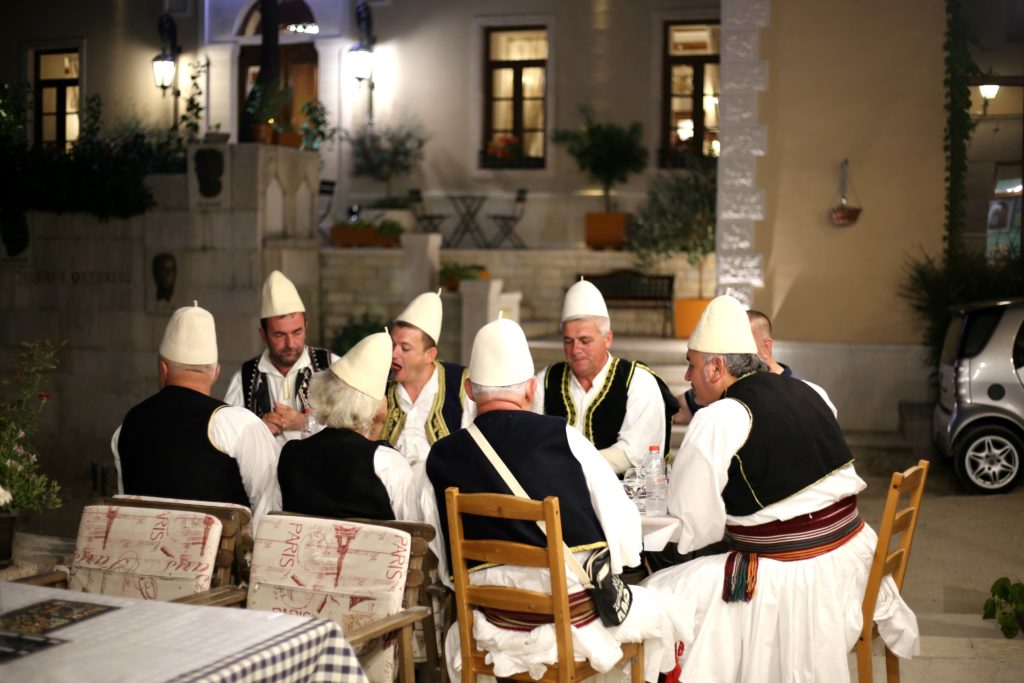 A gathering of men in traditional dress at a table, partaking in the evening xhiro and connecting with neighbors.