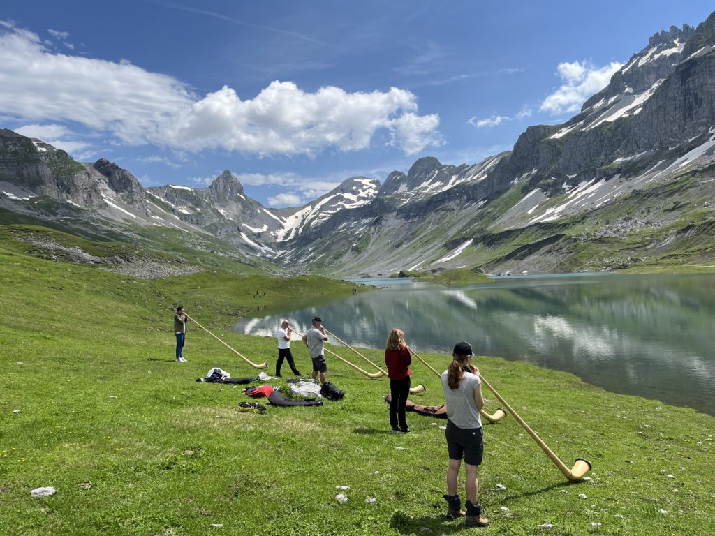 A group of people in the mountains, listening to theoretical instructions in an alphorn workshop.