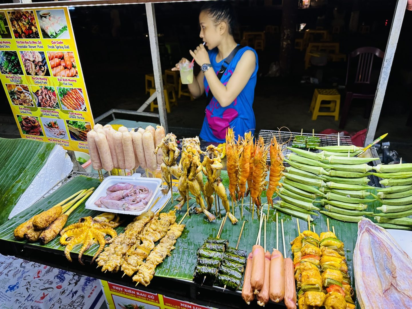 A woman stands at a lively food stand displaying colorful street food offerings