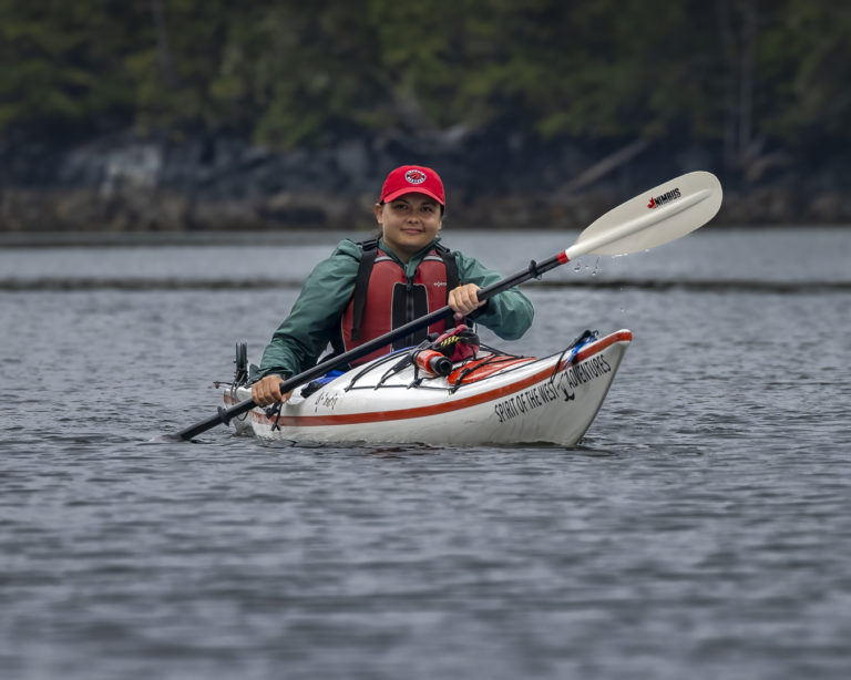 A woman stands near several kayaks on a remote beach, capturing the spirit of sea kayaking