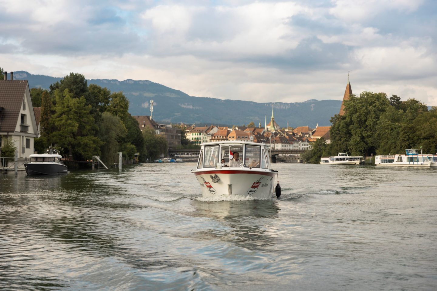 The Öufi boat cruising along the River Aare towards Solothurn’s old town, with church spires and rooftops set against the Jura hills.