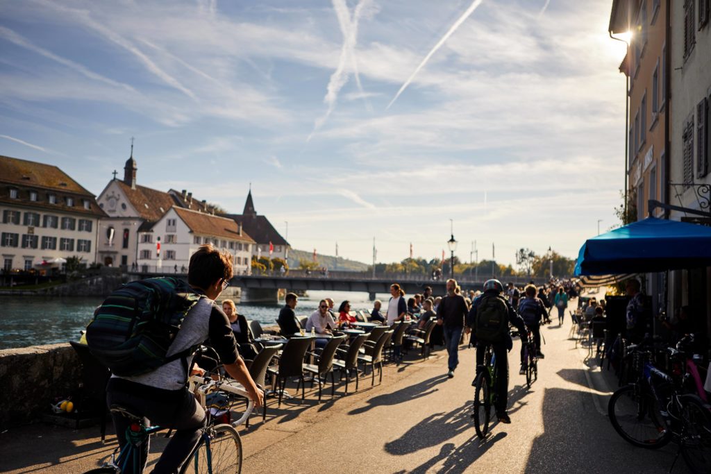 People enjoying a sunny afternoon by the Aare River in Solothurn, relaxing at vibrant riverside cafés and strolling or cycling along the promenade.