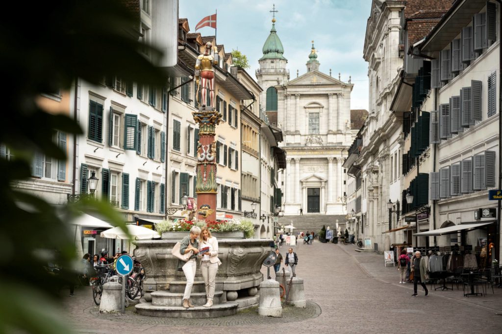 Solothurn’s old town, with two women reading a map beside an ornate fountain and the grand St Ursus Cathedral rising in the background.