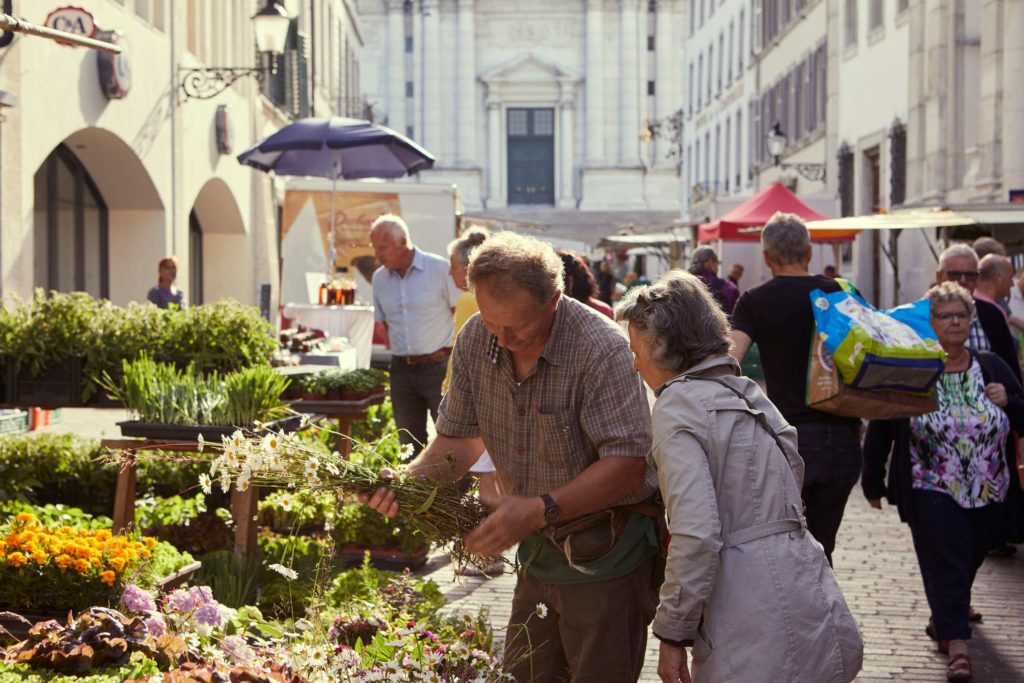 Bustling flower and produce market in Solothurn, with a vendor handing daisies to an older woman, surrounded by colourful plants