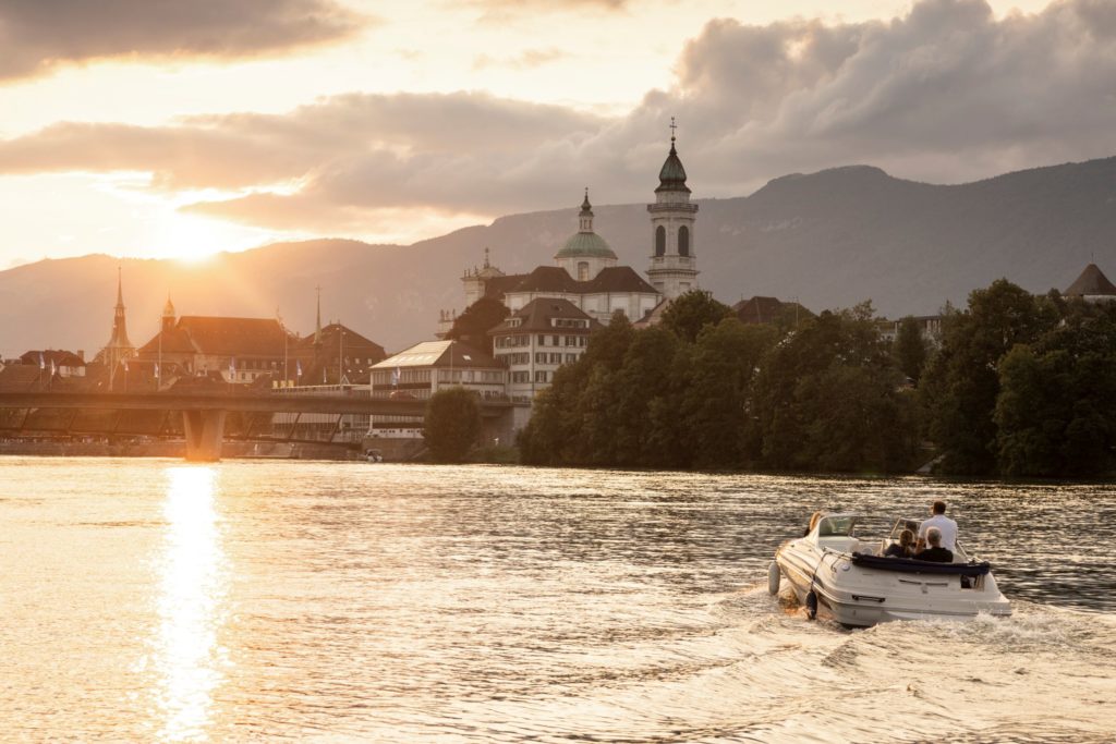 A small motorboat cruising along the River Aare at sunset, with golden light reflecting off the water and the skyline of Solothurn in the background