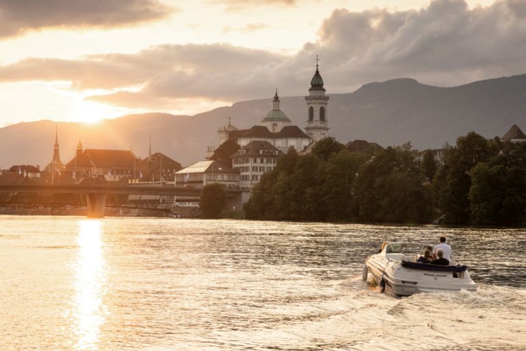 A small motorboat cruising along the River Aare at sunset, with golden light reflecting off the water and the skyline of Solothurn in the background
