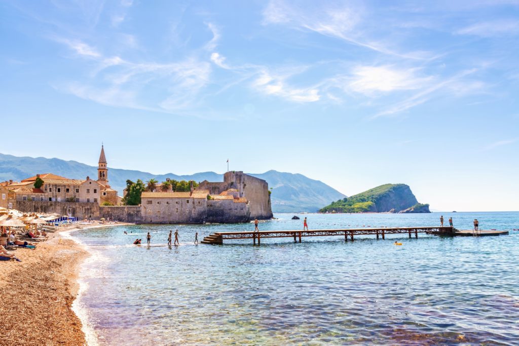 Beachgoers and swimmers by the medieval old town walls of Budva, Montenegro, with clear waters and mountain views.