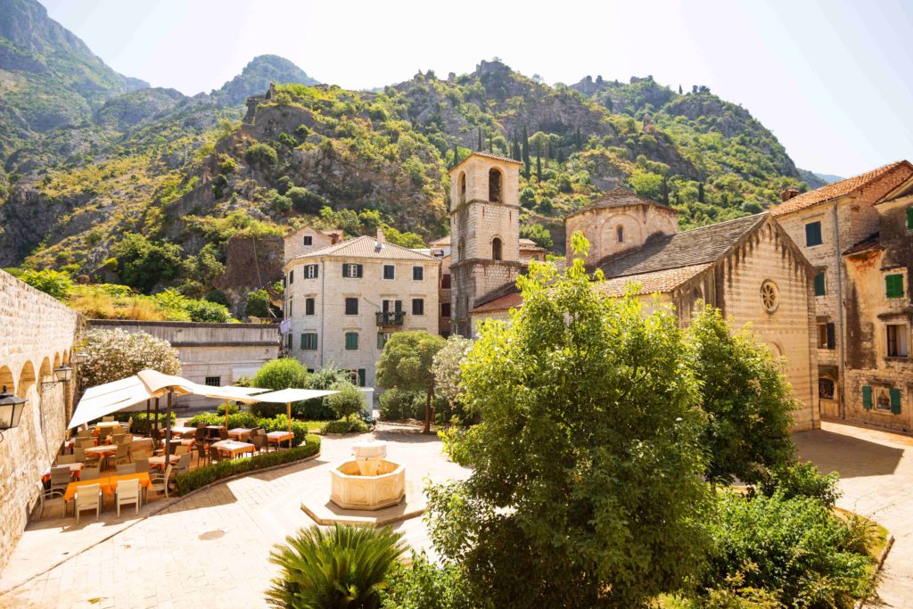 Sunlit square with a church, fountain, and outdoor café in Kotor’s Old Town, Montenegro, backed by green mountains.