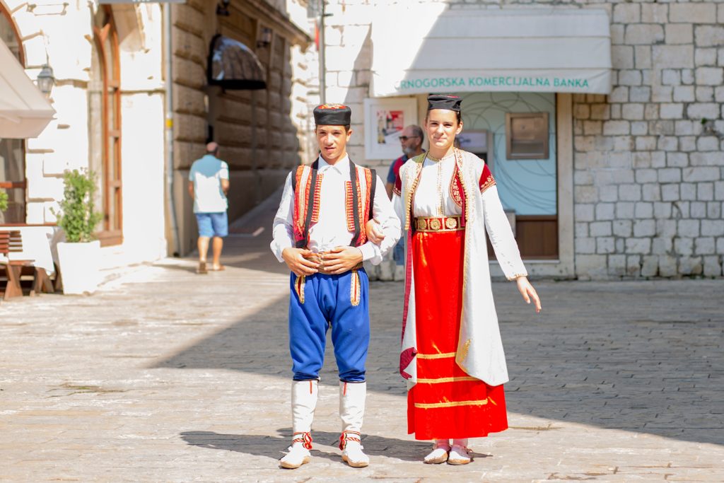 Young man and woman in traditional Montenegrin folk dress standing in a sunny stone-paved square.