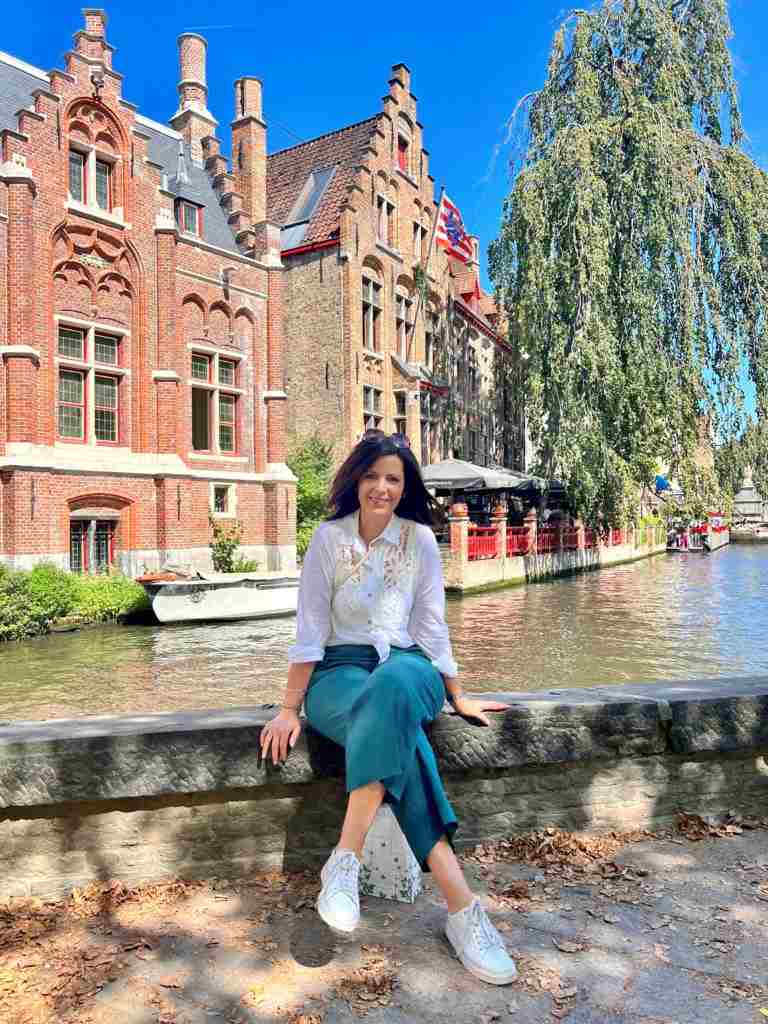Nadia Angelucci sits near a canal in Bruges, with charming buildings.