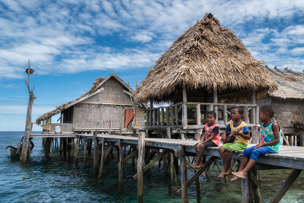Three children from a Papuan island fishing village, sitting on traditional stilt houses built over the water