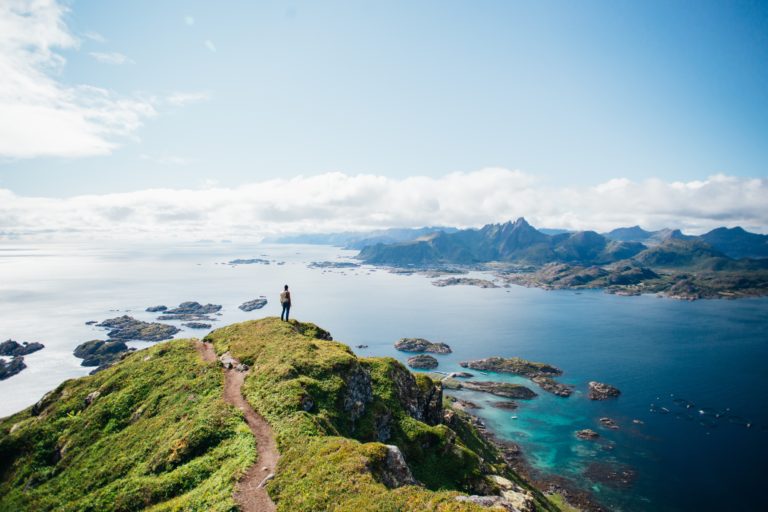 Hiker stands on a mountaintop, overlooking a coastal landscape of islands and fjords in the Lofoten Islands