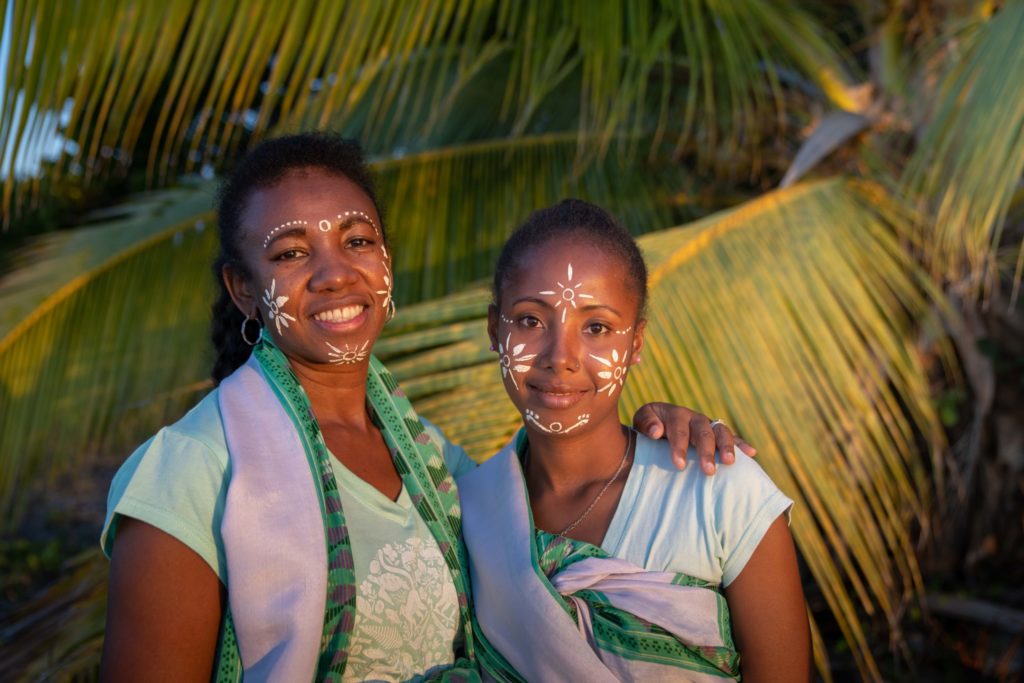 Two women in Madagascar smiling beneath palm leaves, wearing traditional face paint and colourful clothing